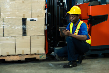 African male inventory worker sitting near wrapped boxes using digital tablet to manage warehouse stock beside forklift in industrial storage building, inside logistics warehouse.