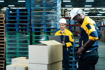 Two multicultural adult male warehouse staff inspecting packages and verifying inventory using laptop surrounded by pallets and boxes in industrial shipping environment, inside logistics warehouse.