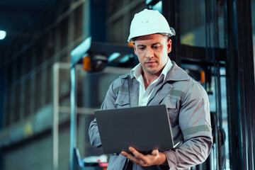 Caucasian male supervisor wearing safety helmet and gray workwear using laptop near forklift...