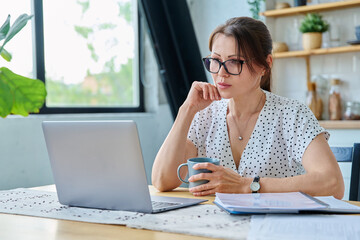 Middle aged serious woman working with laptop sitting at table at home