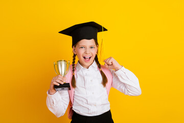 Excited blonde schoolgirl with braids celebrates success holding golden trophy on vibrant yellow...