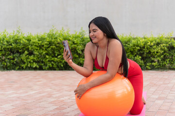 Latin woman doing pilates exercises with a fitness ball and watching online tutorials on smartphone