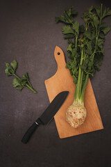 Celery  roots with green stems on a wooden cutting board with black knife on a dark background, top...