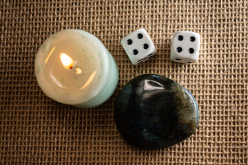 A lit candle, dice, and labradorite rest on burlap, bathed in soft pink light. A mystical flat lay...