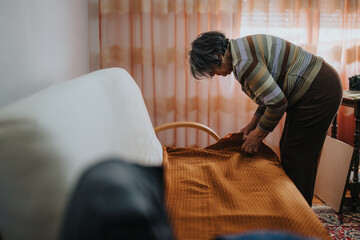 An elderly woman involved in household activity, tidying up and placing a blanket in a warmly lit...