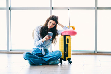 Charming woman sitting on floor with luggage and phone, waiting for flight at airport, feeling...