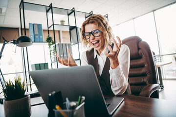 Confident woman expressing excitement while working on a laptop in a modern office environment, productive and happy