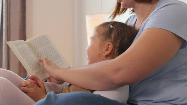 Loving mom reads aloud while lying with her child in a bedroom setting. Childhood development and language growth in a calm family environment. Side view.