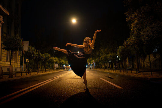 Young ballet dancer in black tutu executing perfect arabesque pose on empty city street illuminated by moonlight and street lamps, creating dramatic nighttime performance