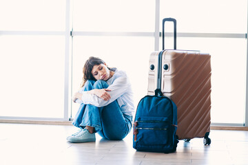 Young woman sitting on floor at airport terminal with luggage, expressing fatigue during travel