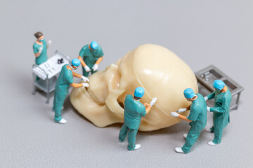 Tiny toy doctors in scrubs perform a delicate and precise surgery on a large human skull model,...