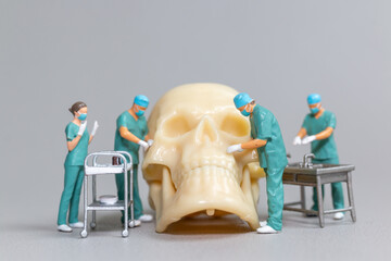Tiny toy doctors in scrubs perform a delicate and precise surgery on a large human skull model,...