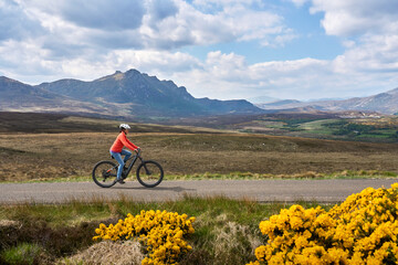 nice senior woman riding her electric mountain bike between blooming gorse in the highlands at the...