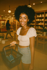 Smiling young woman shopping in store holding basket with products wearing crop top and denim shorts in warm lighting