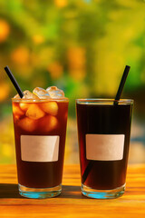 Two glasses of iced and cold brew coffee with straws on wooden table outdoors with sunny green blurred background