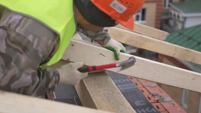 Sesoned builder skillfully drives nail into thick timber beam with practiced force. Wooden framework takes shape under bright natural light