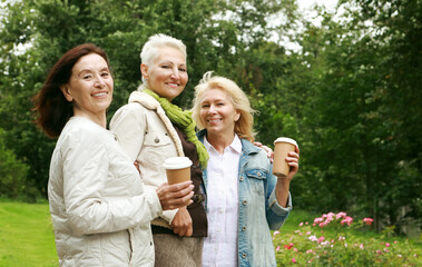 Three cheerful pensioners are walking in the park, having fun talking and drinking coffee.
