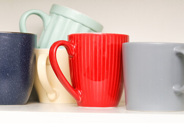 Colorful ceramic mugs on white shelf. Assorted kitchen cups in different shapes and colors. Red,...