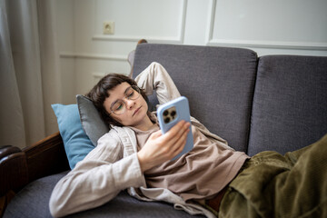 Lazy woman browsing social media on smartphone while lounging on sofa, absorbed in digital web...