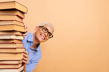 Elderly woman peeking around a stack of book against a beige background, wearing glasses and a...