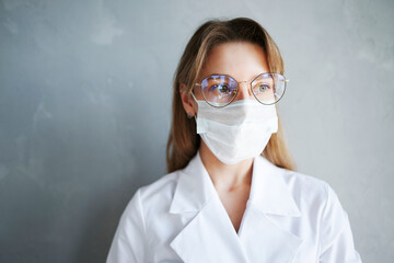 Young female doctor in a medical face mask and eyeglasses looking aside, wearing a white coat against gray background.