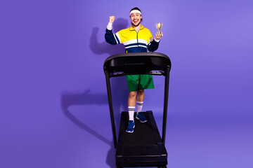 Retro styled young man on treadmill holding trophy, wearing vintage activewear and headband,...