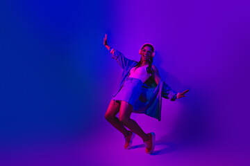 Lovely Young woman with stylish denim outfit and headphones dancing in vibrant neon light