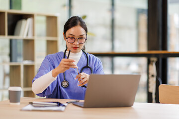 Female medical intern in blue medical scrubs e-learning on laptop computer at the ordination,...