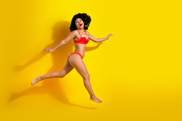 Young woman in vibrant red swimsuit jumping joyfully against yellow background, expressing happiness and summer vibes