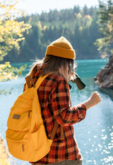 Young woman wearing yellow hat, backpack and plaid shirt stands by serene lake with binoculars...