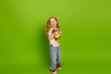 Cute young girl in casual attire standing against a green background, holding a stuffed toy and...
