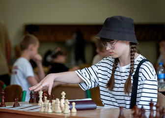 A young woman in glasses concentrates on a move while playing chess. A girl plays in a chess tournament.  Girl at the chessboard. Tournament, portrait of teenage girl playing chess	