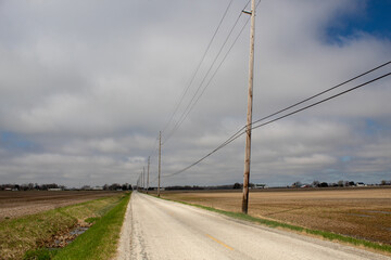 A road along the countryside with farm land on both sides of the road. View of utility poles.