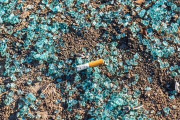 Broken car glass and a cigarette butt on the ground. Blue-green shards on dry soil, symbol of...