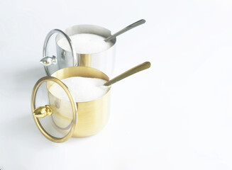 A pair of elegant golden and silver sugar bowls filled with granulated sugar, each topped with a transparent lid and accompanied by a matching spoon on a clean white surface. Photo