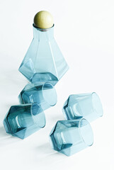A geometric tinted glass decanter with a wooden stopper surrounded by four matching whisky glasses resting on their edges against a bright minimalist background. Photo