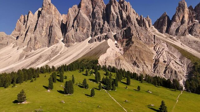 Epic aerial footage of Puez-Odle peaks in the Dolomites, Italy. Jagged ridges rise above alpine meadows and dense spruce forests, glowing under dramatic sunlight
