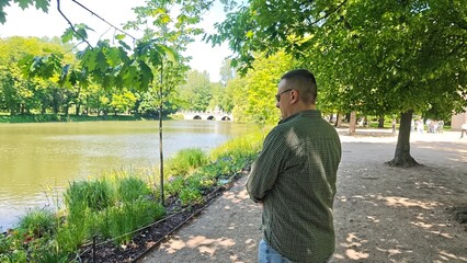 Man standing by a peaceful lake in a sunny park, looking at the water and distant bridge surrounded...