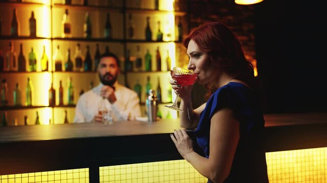 Playful redhead woman sipping cocktail at bar counter, looking around for a date