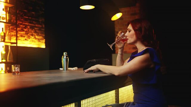 Elegant red-haired woman drinking cocktail at bar counter, searching for a date