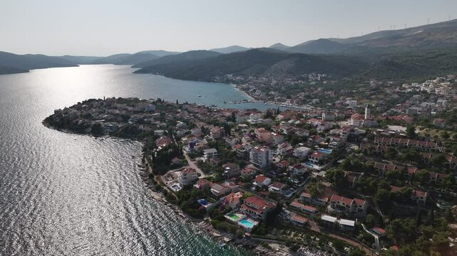 Drone takes a different flight path revealing Seget Vranjica coastline with residential areas, sea and small boats moving near the shore in summer light