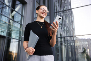 Successful businesswoman with smartphone and folder near office building, low angle view