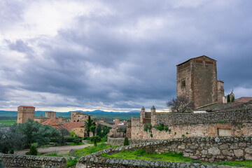 A panoramic view of the historic town of Trujillo Spain, showcasing its ancient stone towers, traditional buildings, and defensive walls under a cloudy sky.