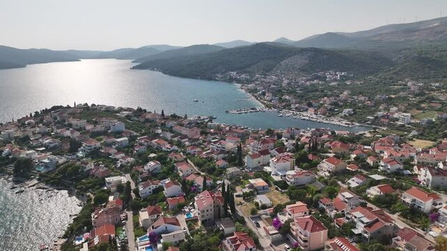 Drone continues to glide above the coastline of Seget Vranjica showing detailed view of the waterfront, harbor, rooftops and boats in motion under soft skies