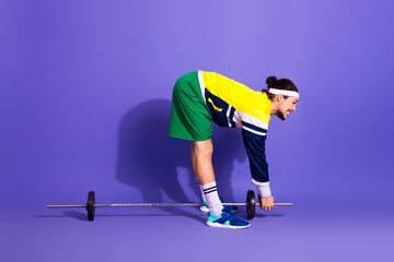 Active athletic man preparing for fitness training with barbell on purple background wearing...