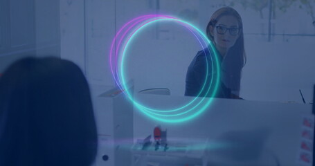 Typing woman wearing glasses, blazer at office desk behind glass partition with holographic overlay