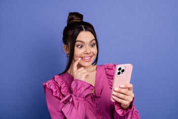 Smiling young woman in a stylish pink dress holding a smartphone against a violet background,...