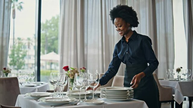 positive female african american waiter serving table with empty plates in restaurant