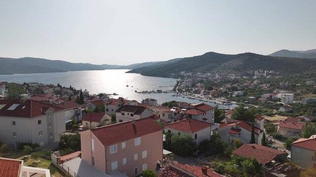 Drone hovers over elevated position above Seget Vranjica showing houses, hills and the shimmering Adriatic Sea during a bright and warm afternoon