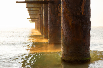 Long pier ruins pointing toward horizon over Black Sea, sun low over water, calmness and decay in balance. Underneath old pier with decaying steel and concrete at sunset, sea motion and golden colors 